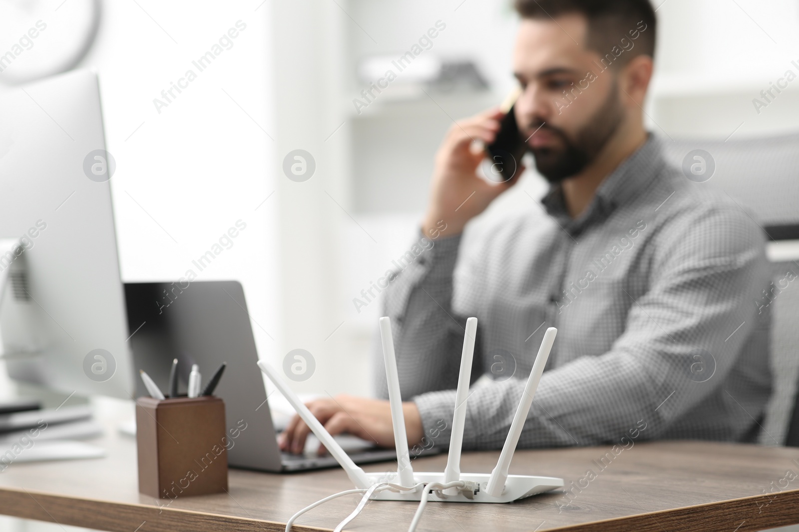 Photo of Man talking on smartphone while working at wooden table indoors, focus on Wi-Fi router