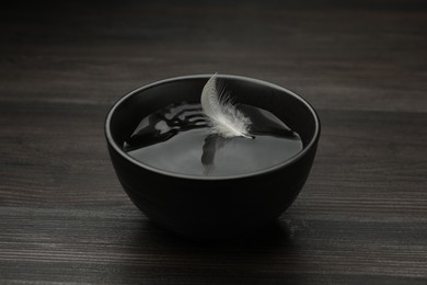 Photo of Black bowl with water and white feather on dark wooden table