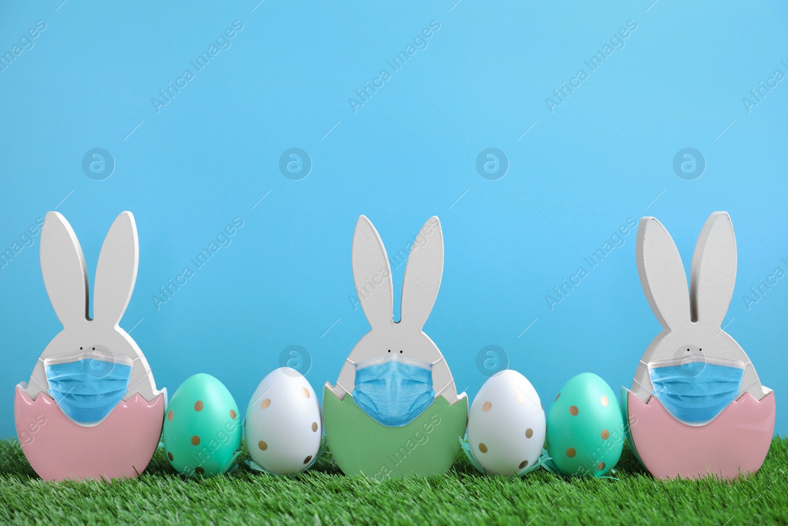 Image of COVID-19 pandemic. Easter bunny figures in protective masks and dyed eggs on green grass against light blue background