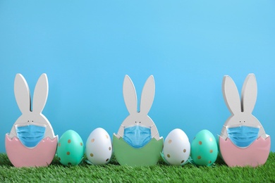 Image of COVID-19 pandemic. Easter bunny figures in protective masks and dyed eggs on green grass against light blue background