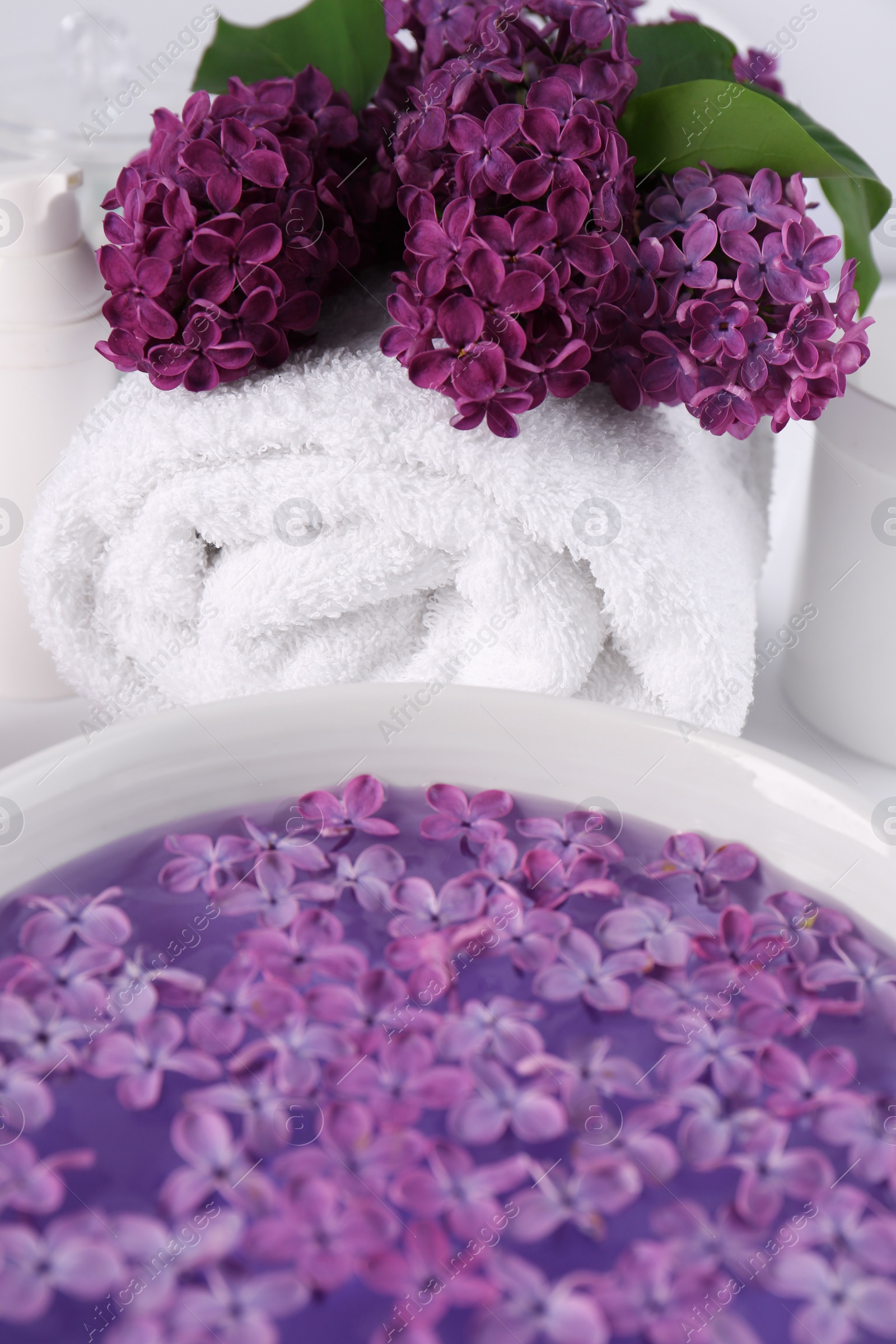 Photo of Lilac flowers with white towel and bowl of aromatic water on table, closeup
