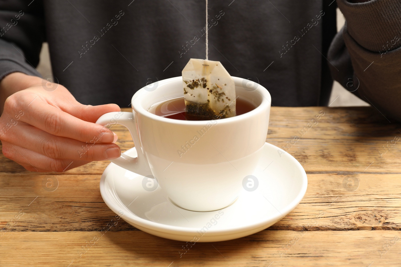 Photo of Woman putting tea bag in cup with hot water at wooden table, closeup