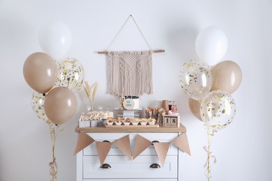 Baby shower party. Different delicious treats on white wooden chest of drawers and decor near light wall