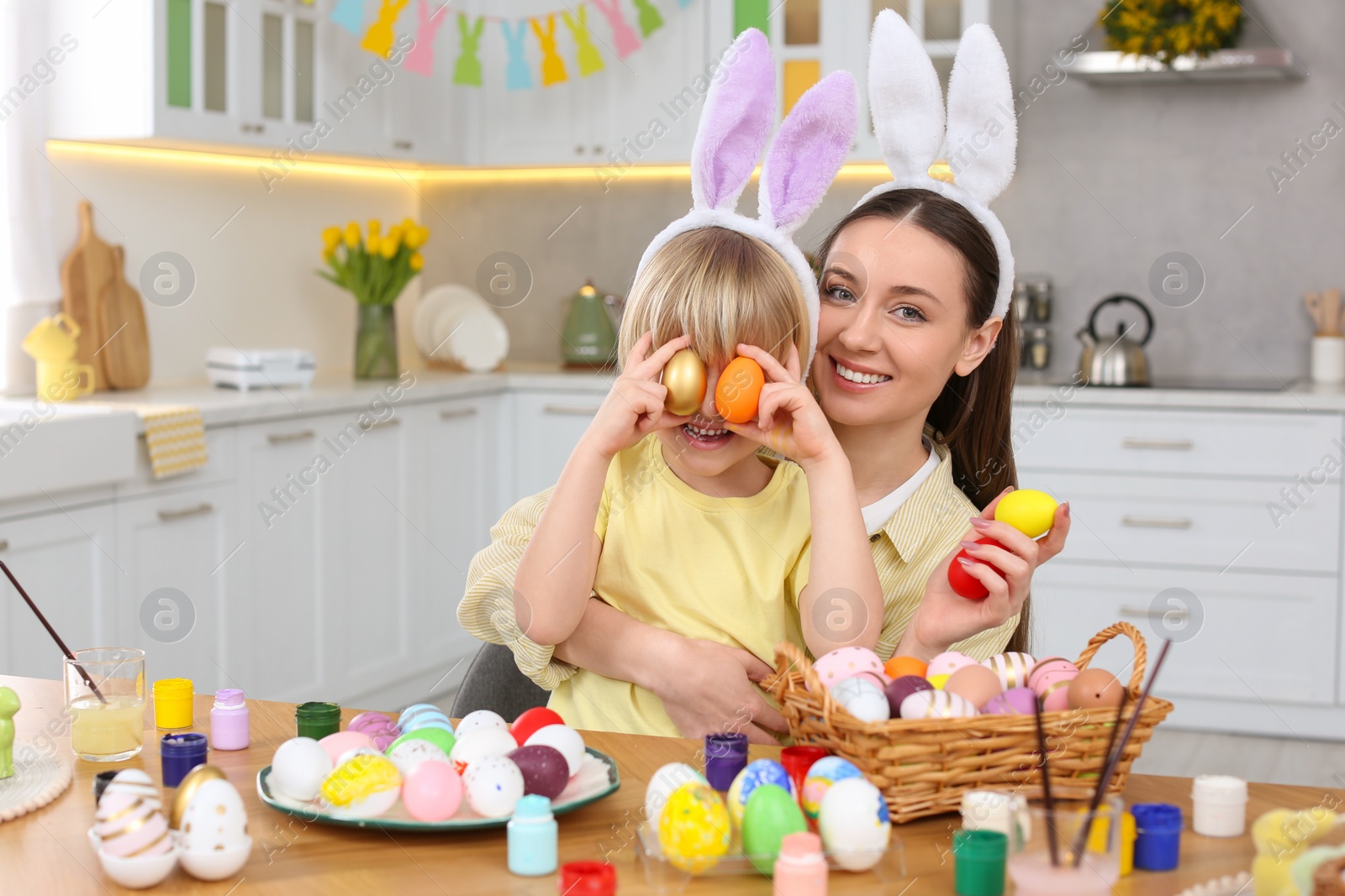 Photo of Mother and her cute son covering eyes with Easter eggs at table in kitchen