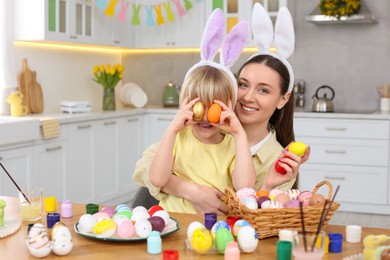 Mother and her cute son covering eyes with Easter eggs at table in kitchen