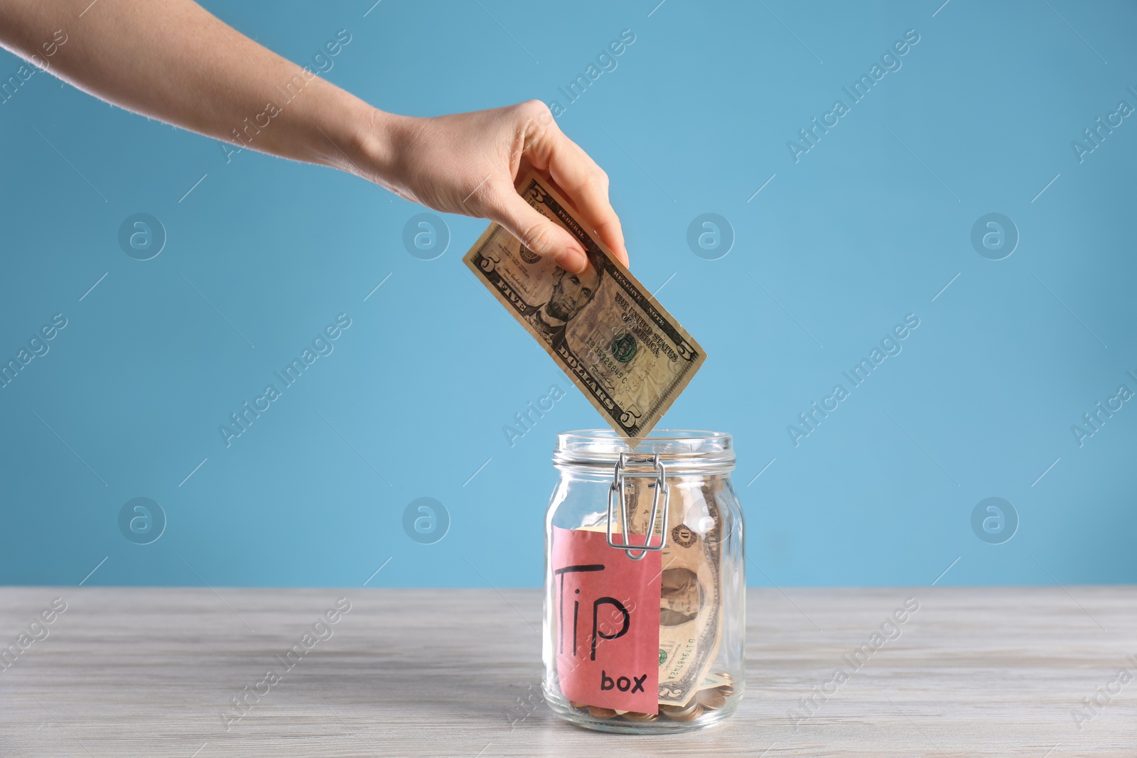 Photo of Woman putting tips into glass jar on wooden table, closeup