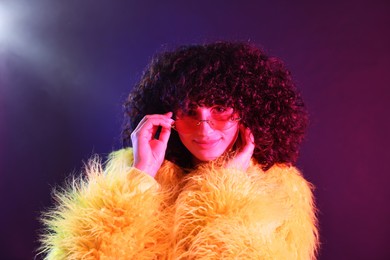 Beautiful young woman in yellow fur coat and sunglasses on color background in neon lights