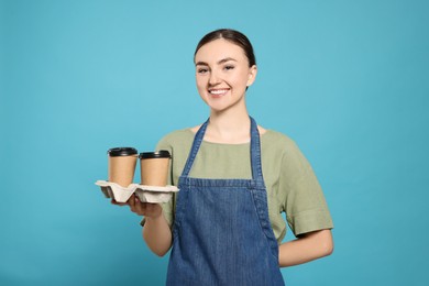 Beautiful young woman in denim apron with delicious coffee on light blue background