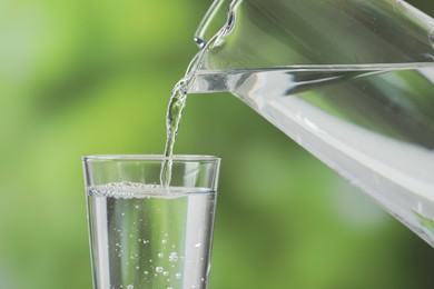 Pouring fresh water from jug into glass against blurred green background, closeup