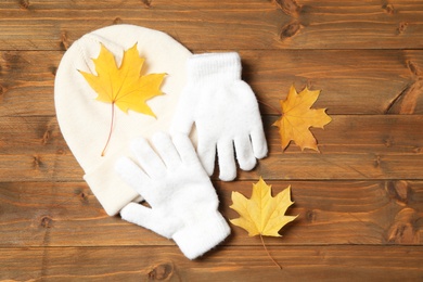 Photo of Stylish white woolen gloves, hat and dry leaves on wooden table, flat lay