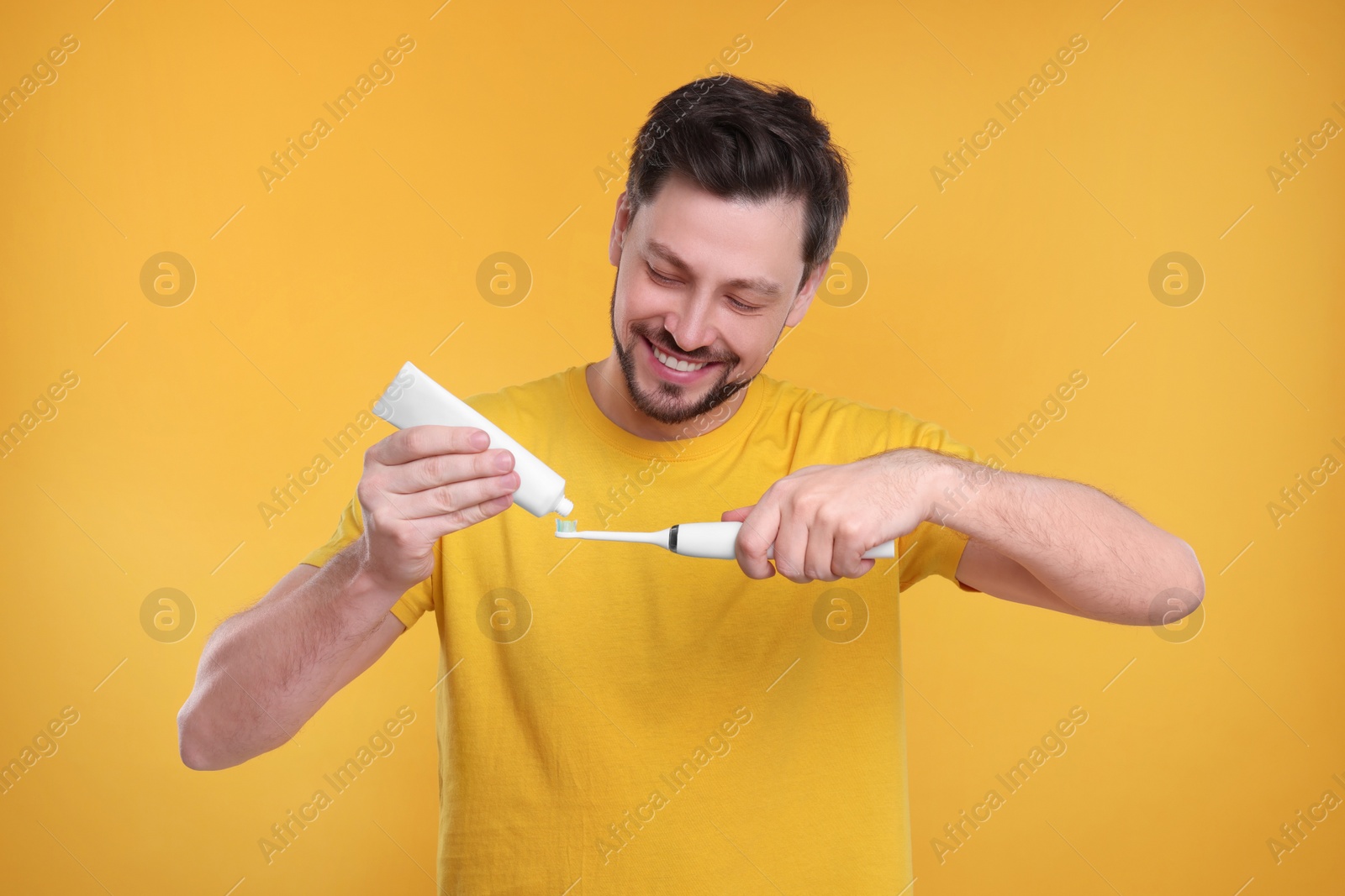 Photo of Happy man squeezing toothpaste from tube onto electric toothbrush on yellow background