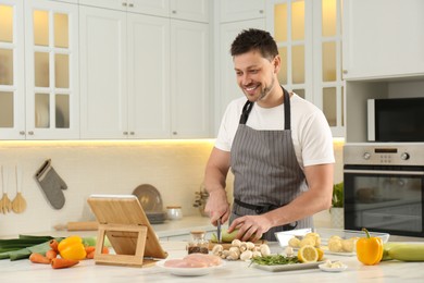 Photo of Happy man making dinner while watching online cooking course via tablet in kitchen