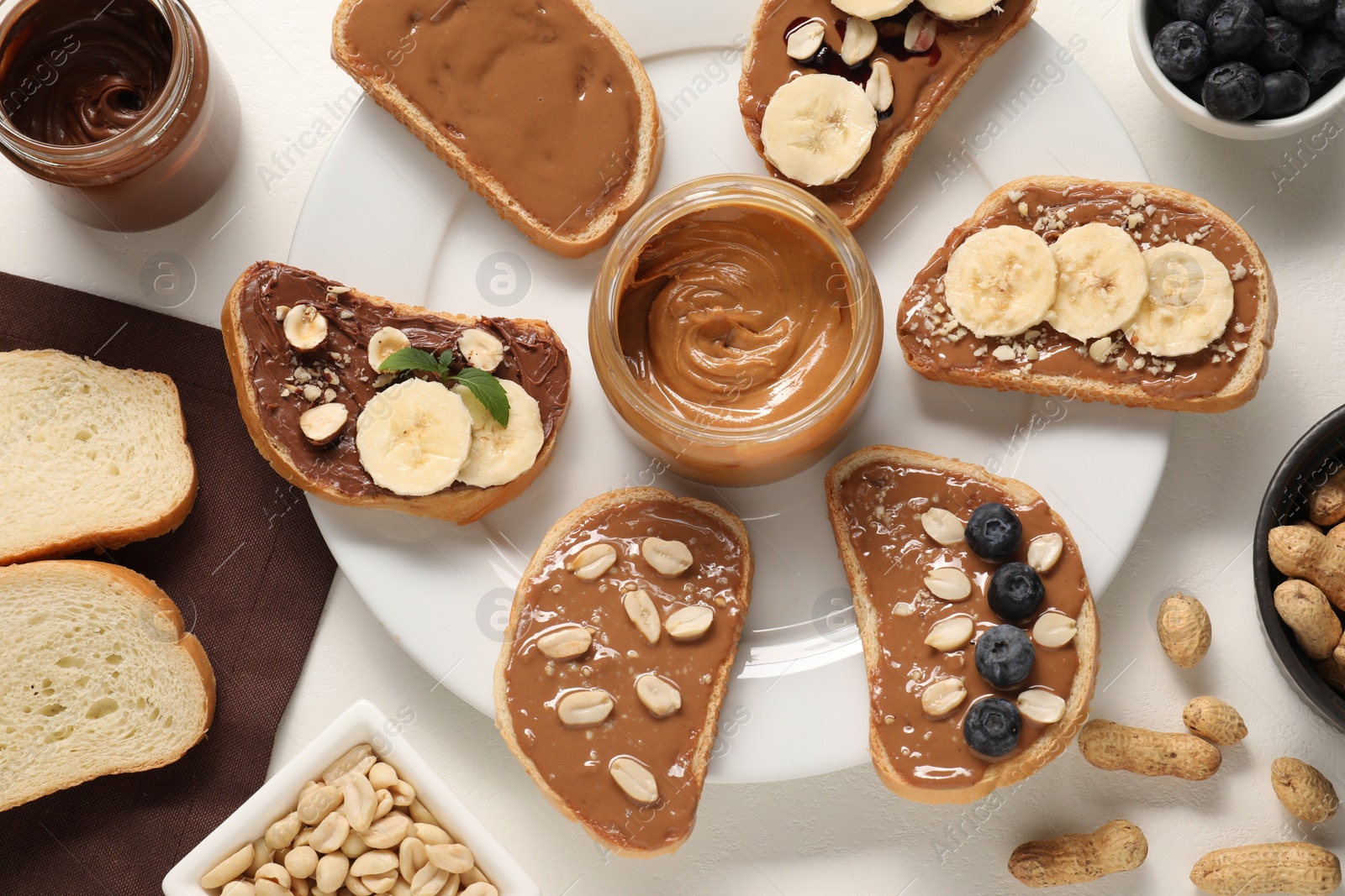 Photo of Toasts with different nut butters, banana slices, blueberries and nuts on white table, flat lay