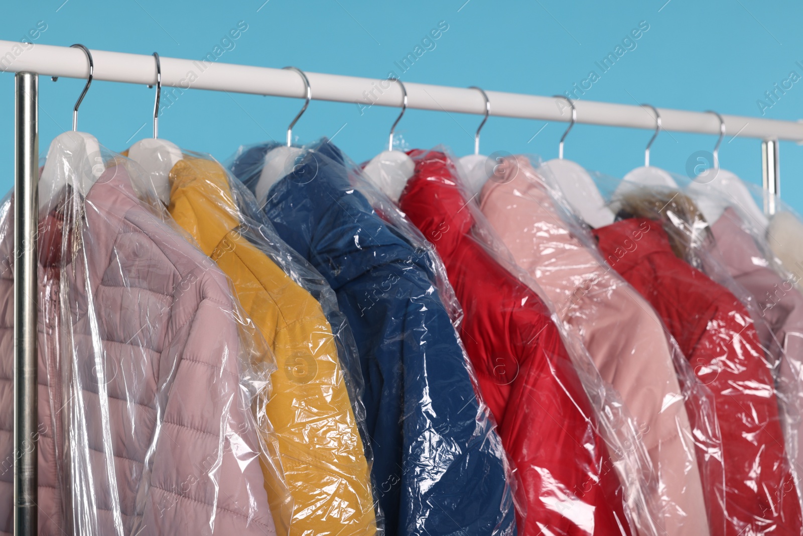 Photo of Dry-cleaning service. Many different clothes in plastic bags hanging on rack against light blue background