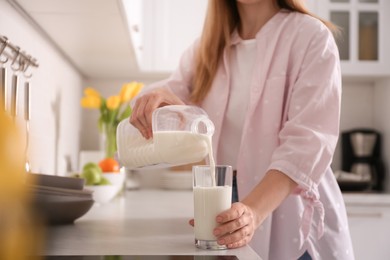 Young woman pouring milk from gallon bottle into glass at light countertop in kitchen, closeup