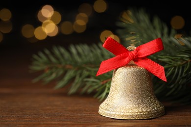 Photo of Bell with red bow and fir branches on wooden table against blurred background, closeup. Christmas decor