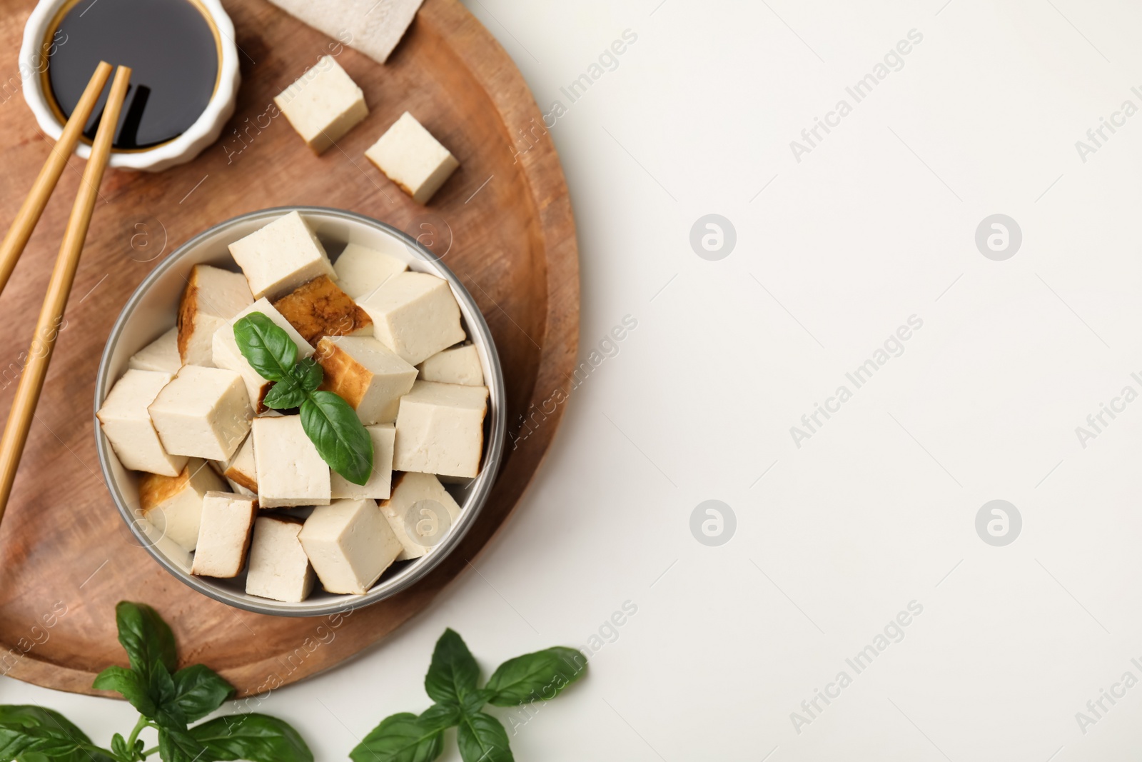 Photo of Wooden tray with bowl of smoked tofu cubes, basil and soy sauce on white table, flat lay. Space for text