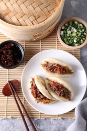 Photo of Delicious gua bao (pork belly buns), chopsticks, green onion and sauce on grey textured table, flat lay