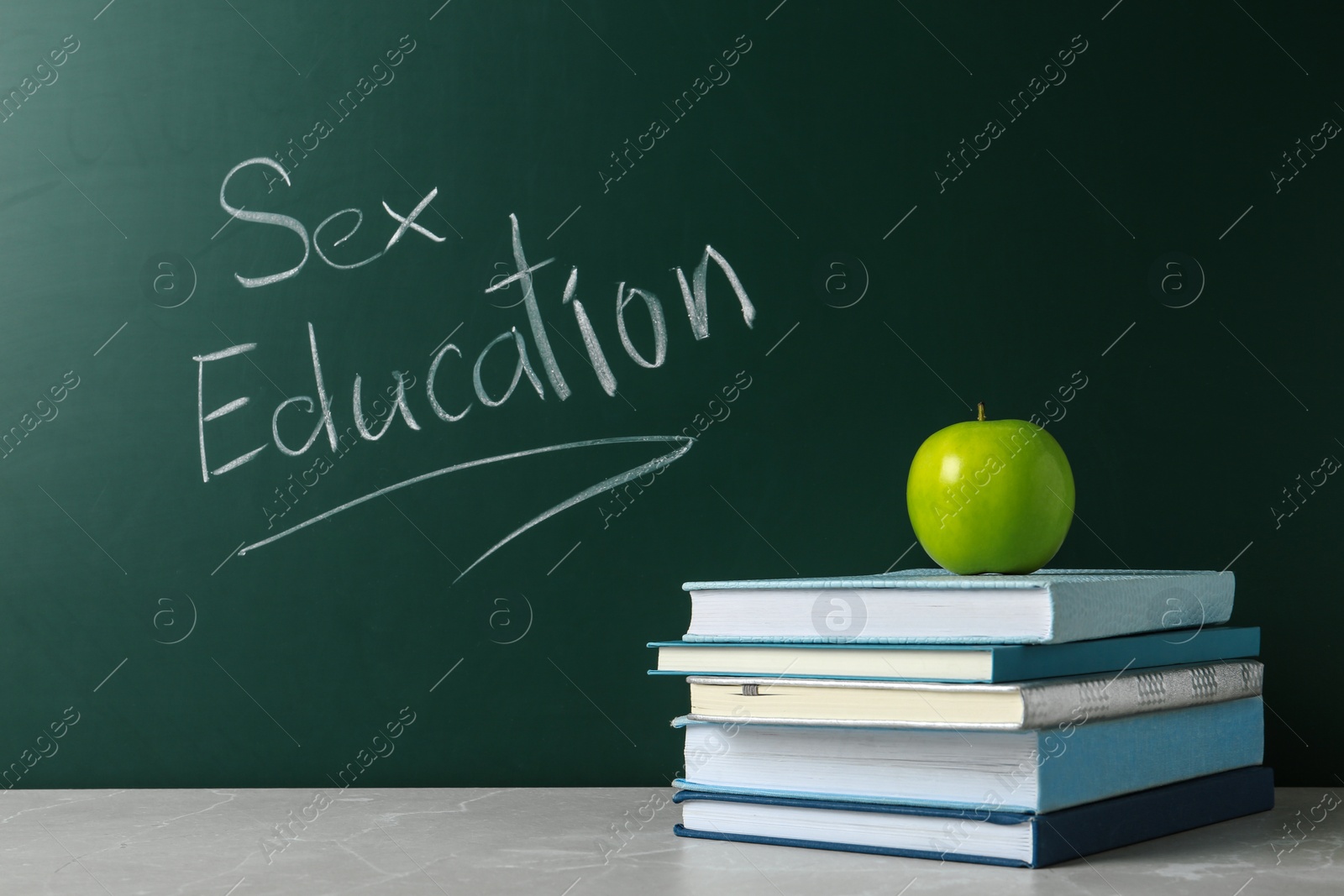 Photo of Books and apple on grey table near chalkboard with phrase "Sex education"