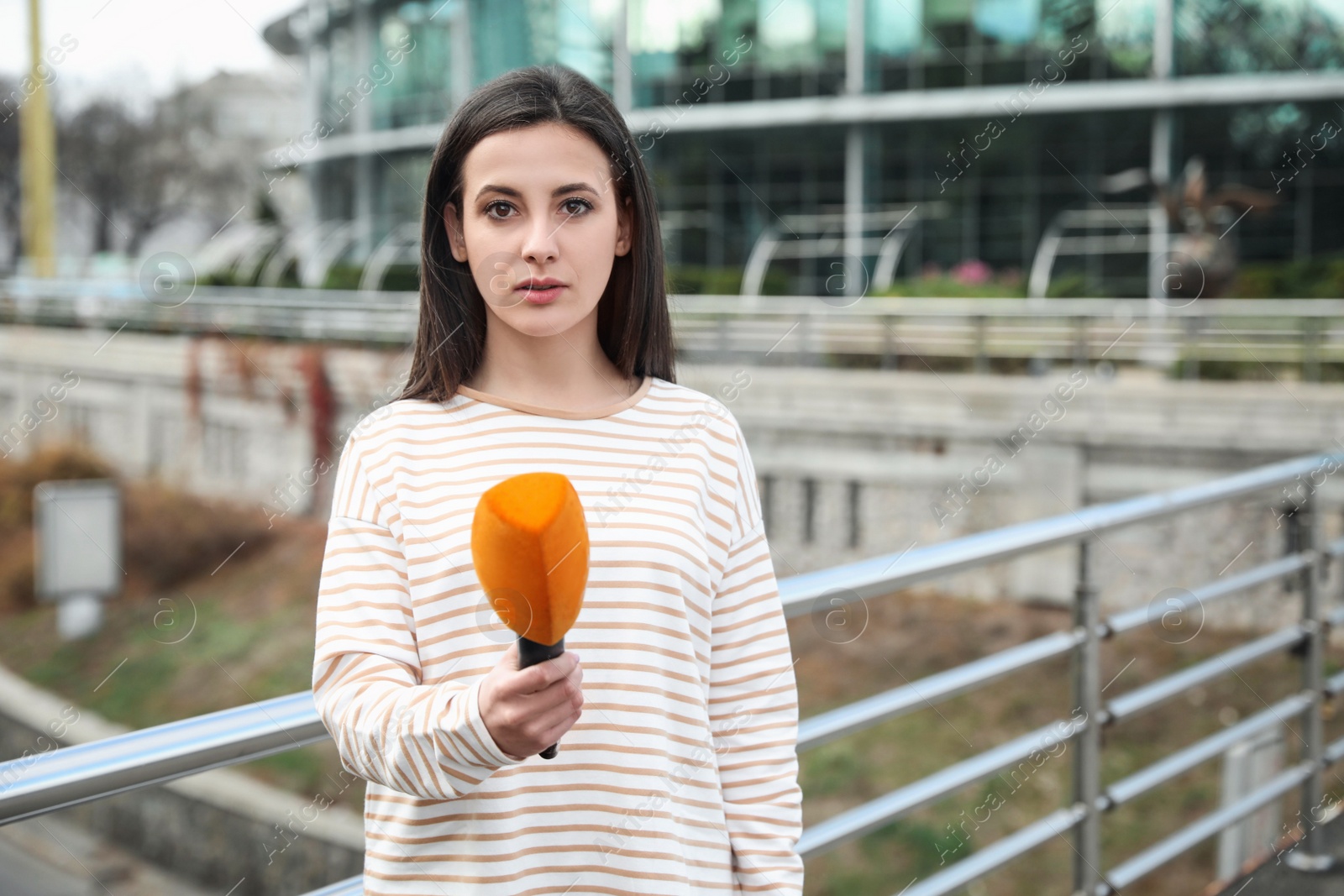 Photo of Young female journalist with microphone working on city street