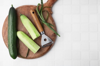 Photo of Fresh cucumbers and peeler on white tiled table, top view. Space for text