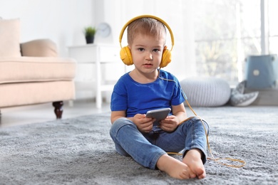 Photo of Cute child with headphones and mobile phone on floor indoors