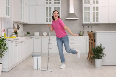 Photo of Enjoying cleaning. Happy woman in headphones dancing with mop in kitchen