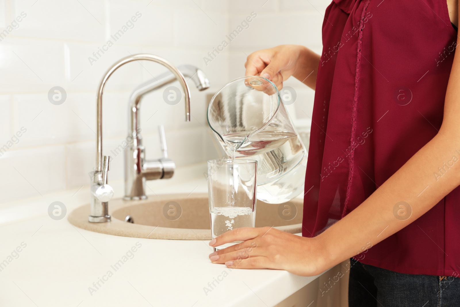 Photo of Woman pouring water from jug into glass in kitchen, closeup