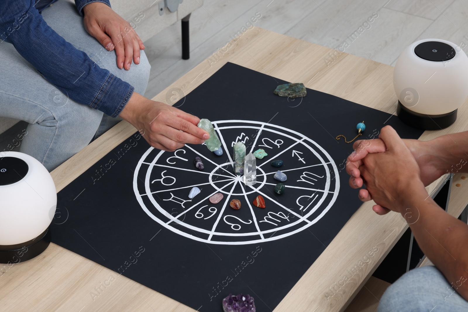 Photo of Astrologer predicting client's future with zodiac wheel, stones and crystals at wooden table indoors, closeup