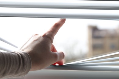 Woman separating slats of white blinds indoors, closeup