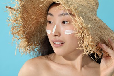 Beautiful young woman in straw hat with sun protection cream on her face against light blue background