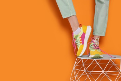 Photo of Woman in stylish colorful sneakers standing on white table against orange background, closeup. Space for text