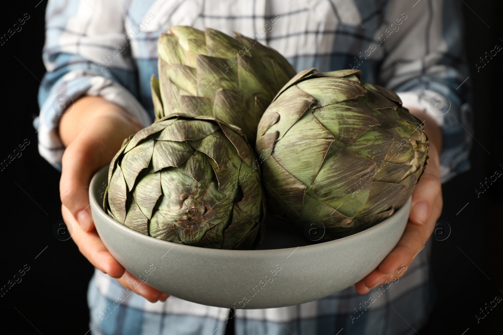 Photo of Woman holding bowl with fresh raw artichokes on black background, closeup