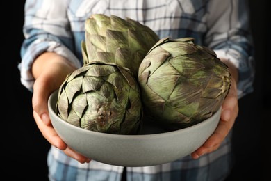 Woman holding bowl with fresh raw artichokes on black background, closeup