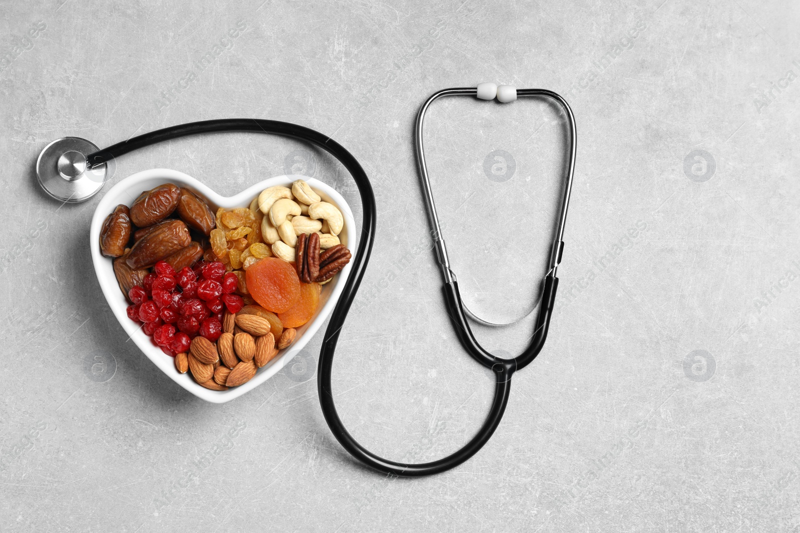 Photo of Heart shaped bowl with nuts and dried fruits near stethoscope on grey background, top view. Space for text