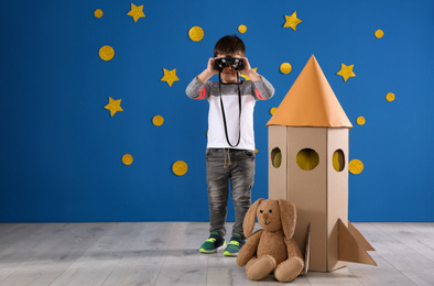 Photo of Cute little child playing with binoculars, cardboard rocket and toy bunny near blue wall