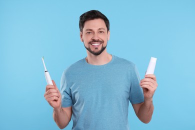 Happy man holding electric toothbrush and tube of toothpaste on light blue background