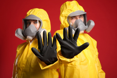 Photo of Man and woman in chemical protective suits making stop gesture against red background, focus on hands. Virus research