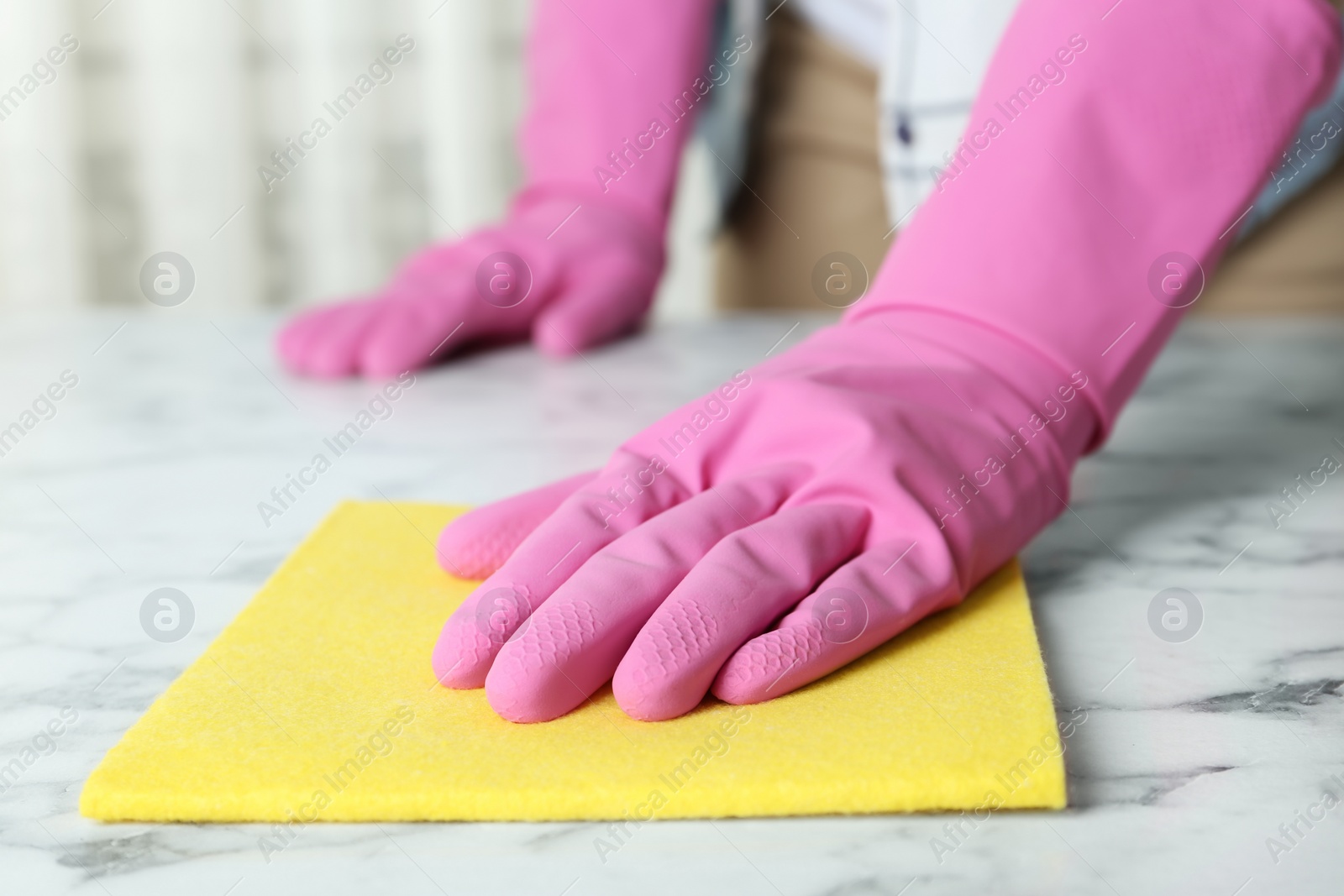 Photo of Woman in gloves wiping white marble table with rag indoors, closeup