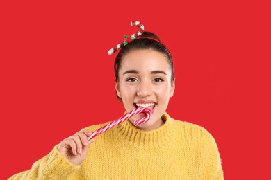 Young woman in yellow sweater and festive headband eating candy cane on red background