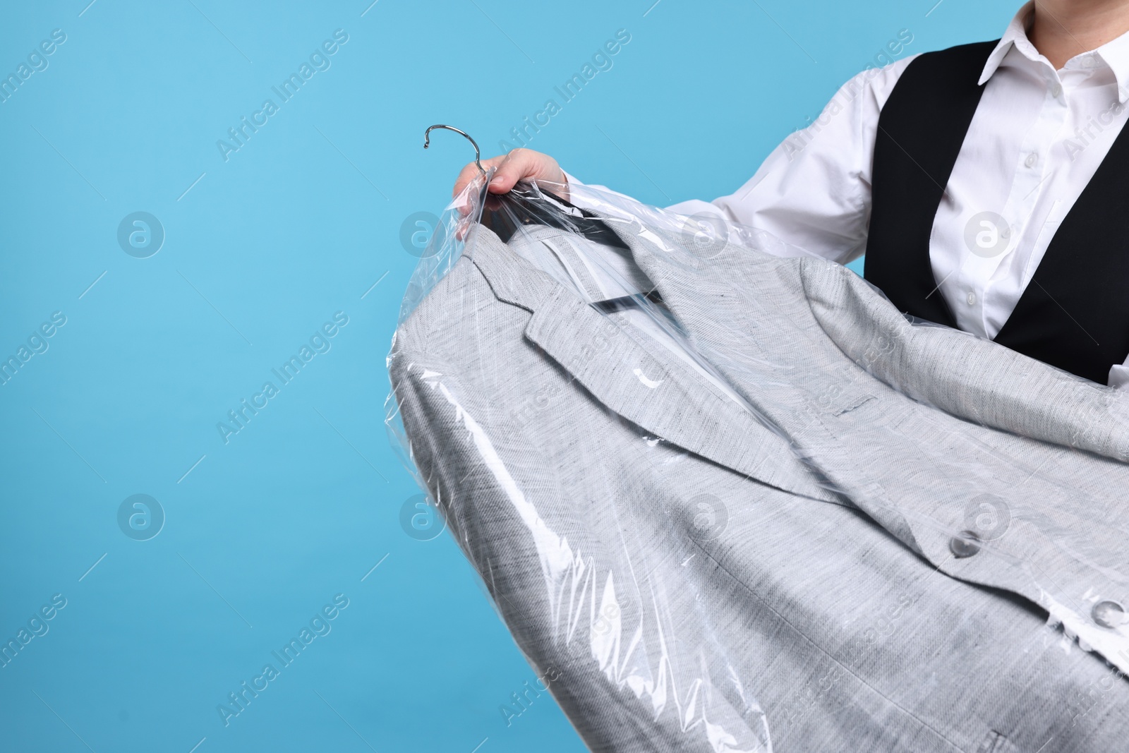 Photo of Dry-cleaning service. Woman holding jacket in plastic bag on light blue background, closeup. Space for text