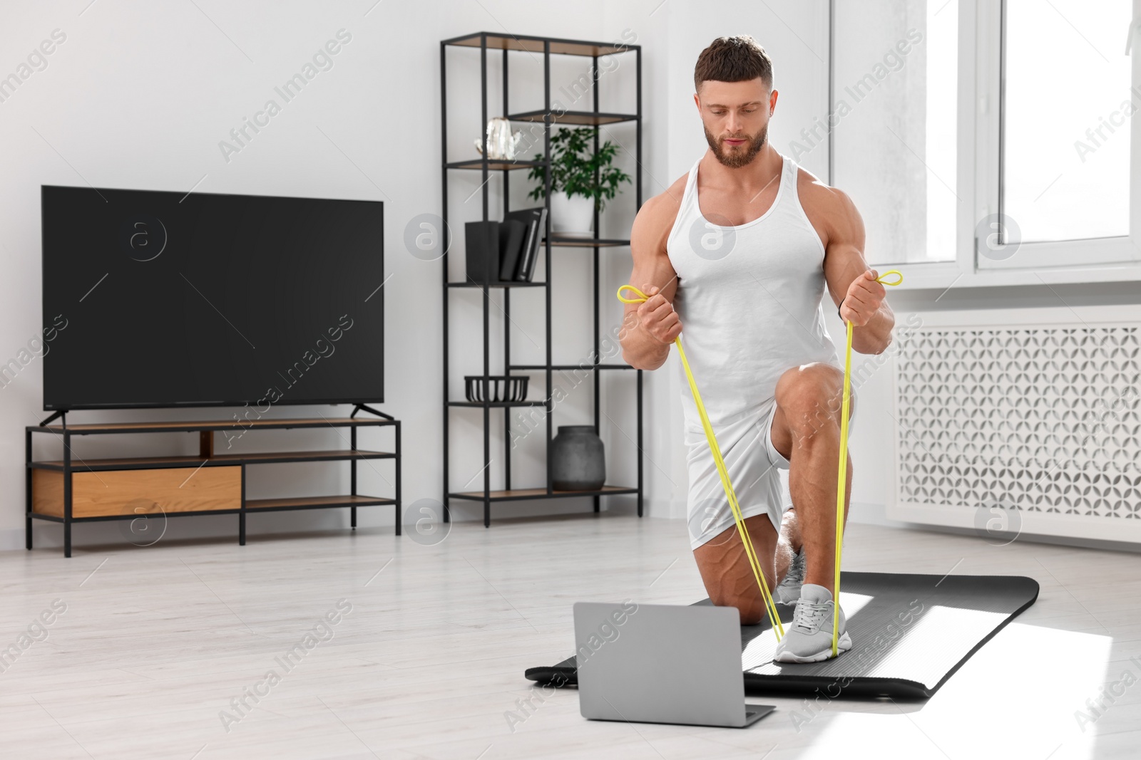 Photo of Muscular man doing exercise with elastic resistance band near laptop on mat at home