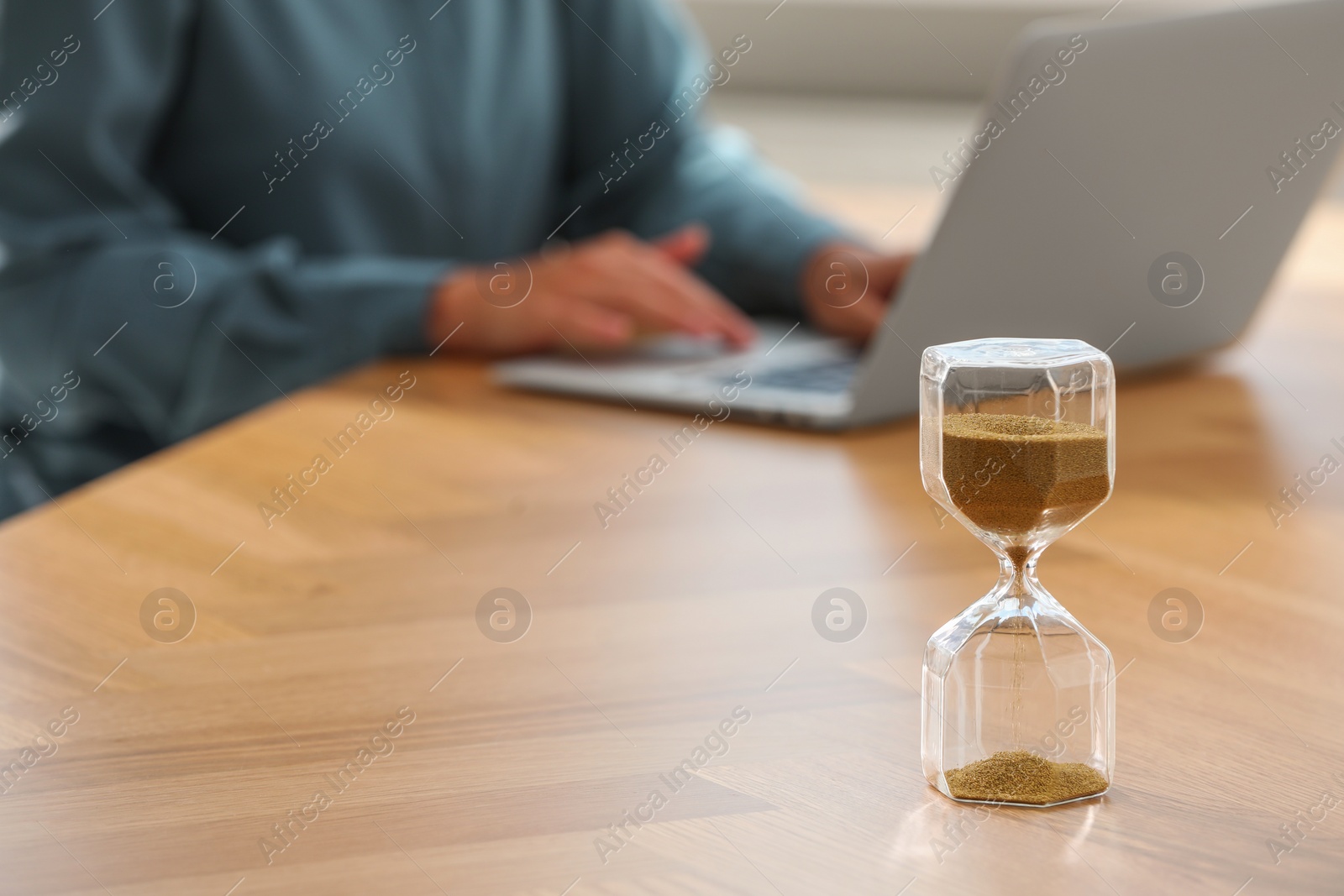 Photo of Hourglass with flowing sand on table. Woman using laptop indoors, selective focus