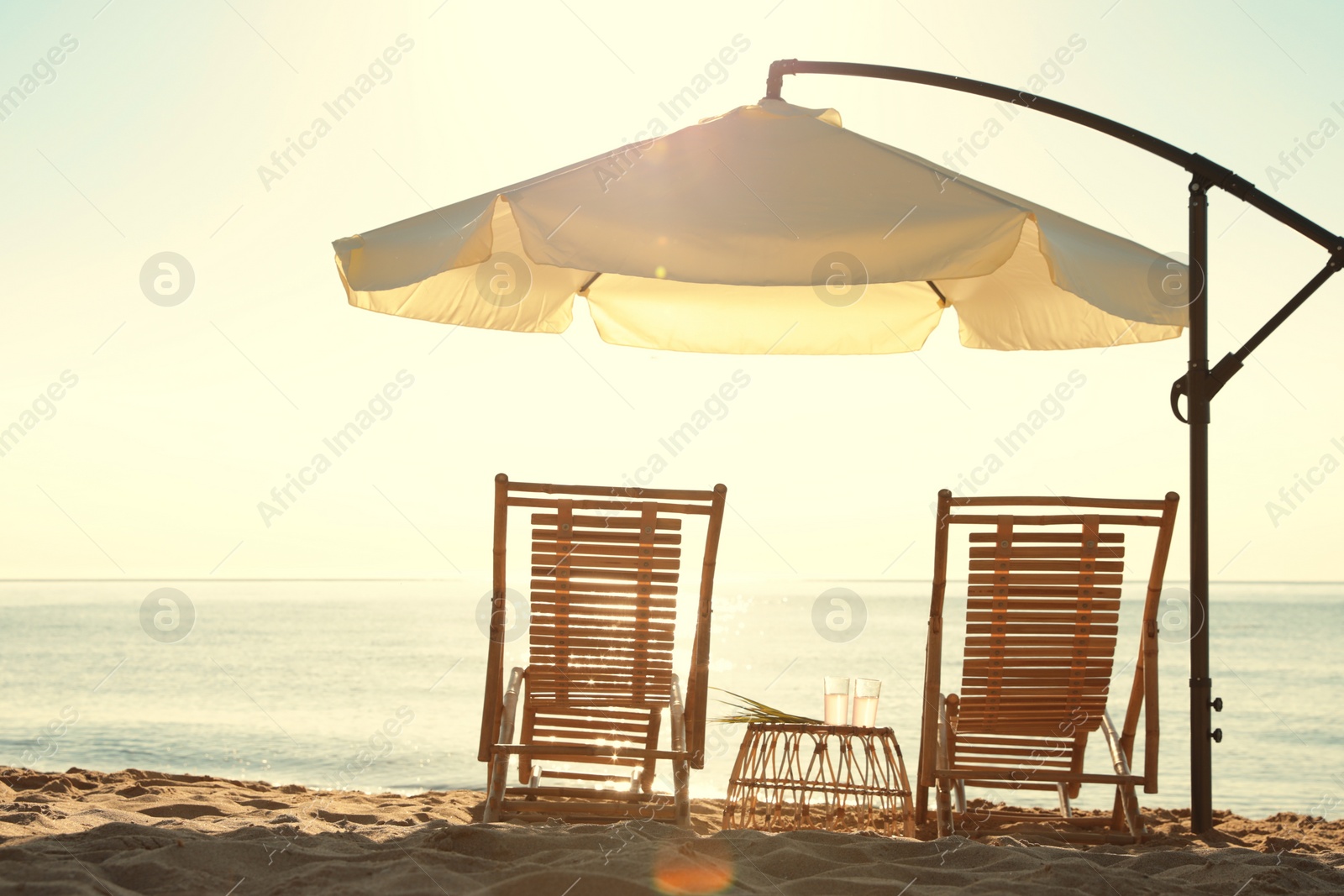 Photo of Wooden deck chairs, outdoor umbrella and table with cocktails on sandy beach. Summer vacation