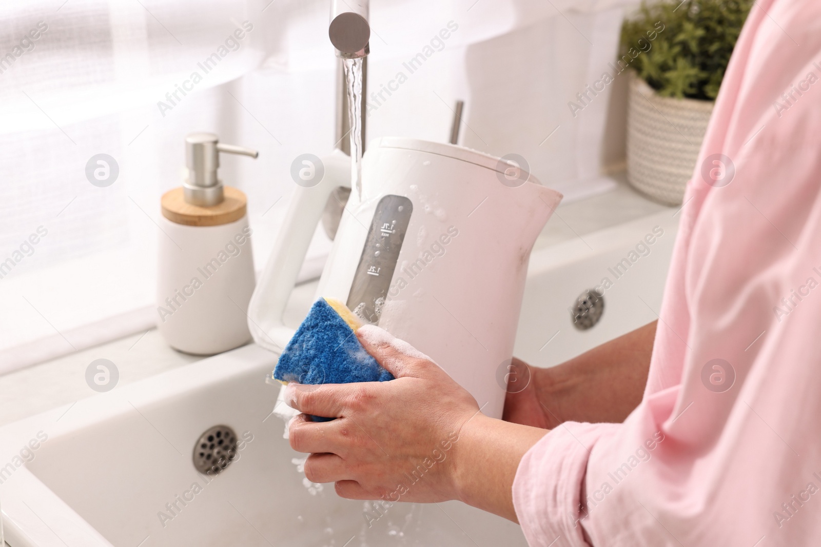 Photo of Woman washing electric kettle with sponge at sink in kitchen, closeup