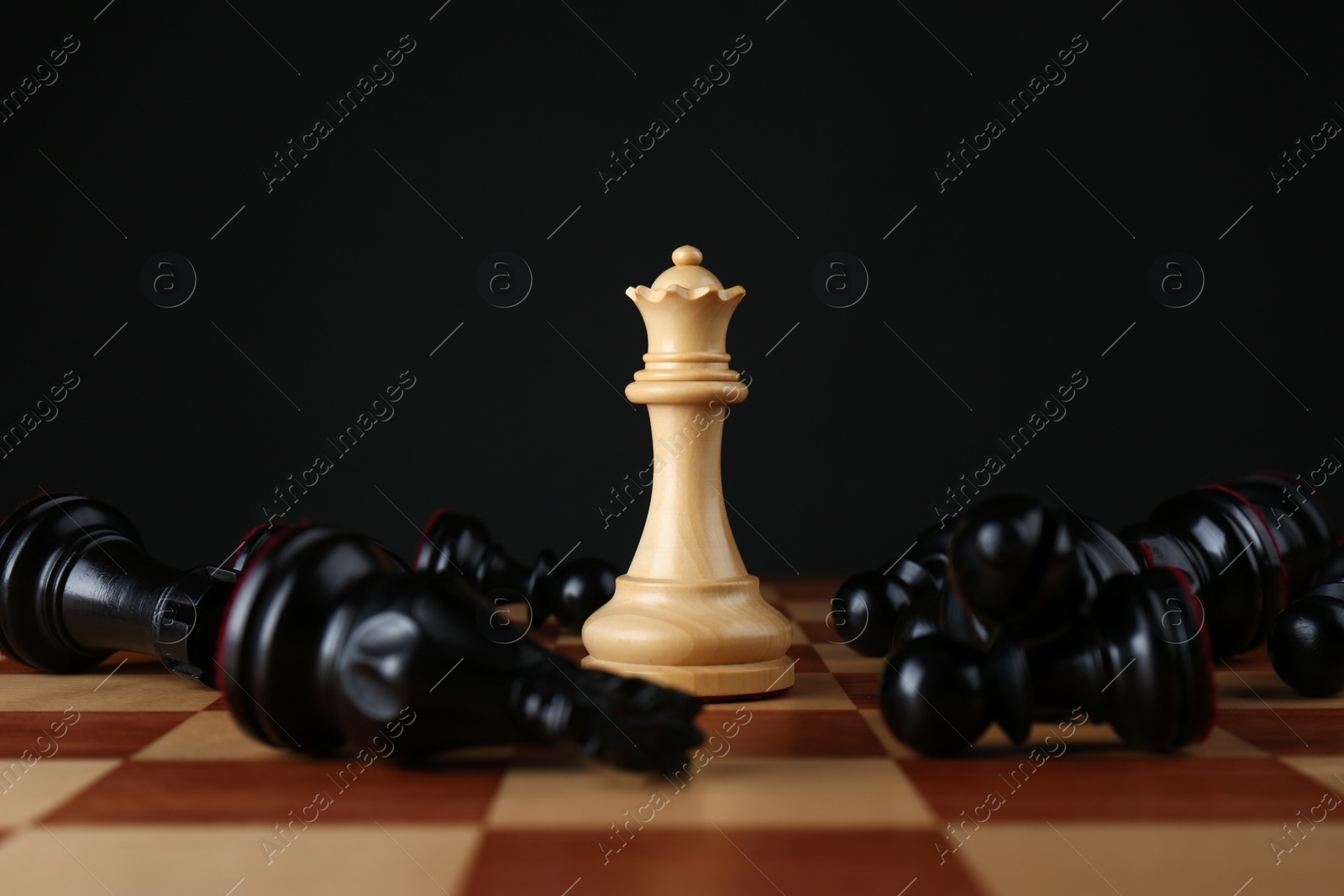 Photo of White queen among fallen black chess pieces on wooden board against dark background