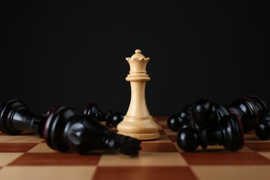 White queen among fallen black chess pieces on wooden board against dark background