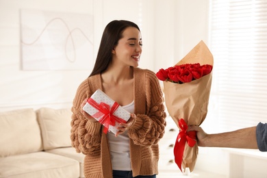 Photo of Happy woman receiving red tulip bouquet and gift box from man at home. 8th of March celebration
