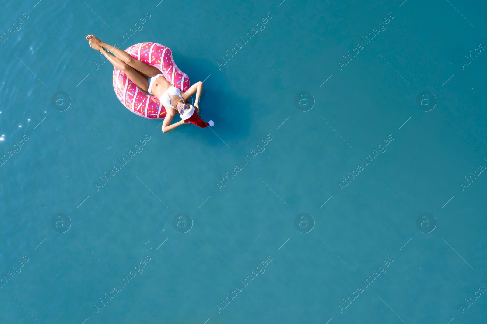 Image of Young woman wearing Santa hat and bikini with inflatable ring in sea, top view. Christmas vacation