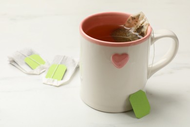 Tea bags and cup of hot beverage on white table, closeup